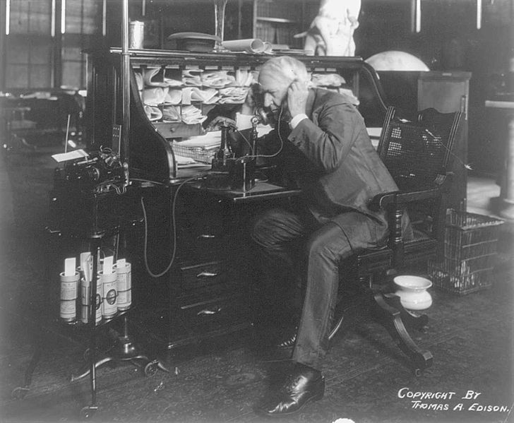 Thomas Alva Edison at his desk, talking on telephone, on 1914 Sept. 8. An Edison wax cylinder dictating machine is seen to the left with several cylinder records beneath it.